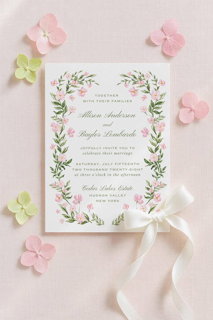 a wedding invitation card with a floral border and a white ribbon bow, surrounded by pink and green flowers on a light pink background.