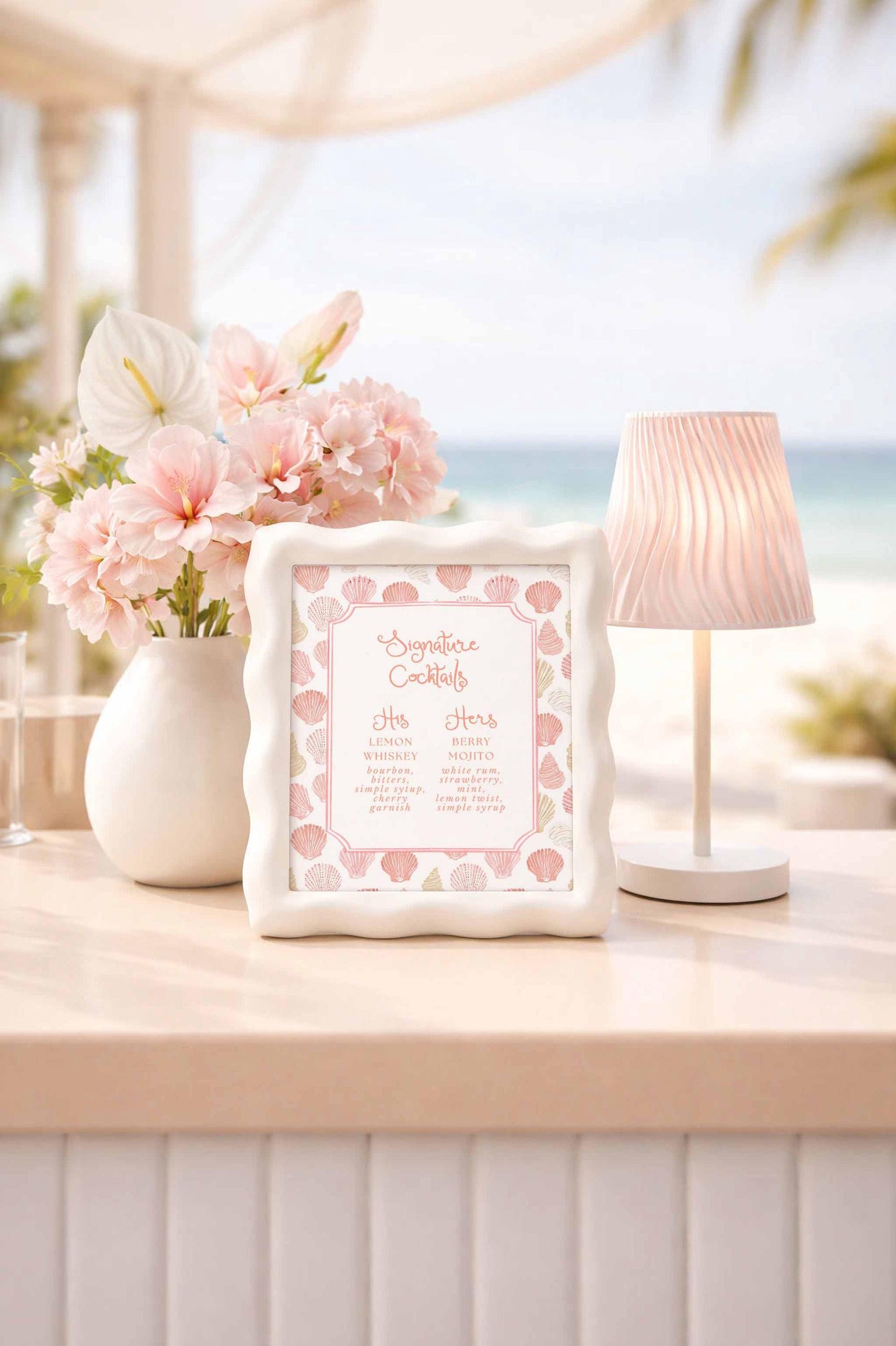 a table with a framed cocktail menu, a vase of pink flowers, and a lamp, all set against a beach background.