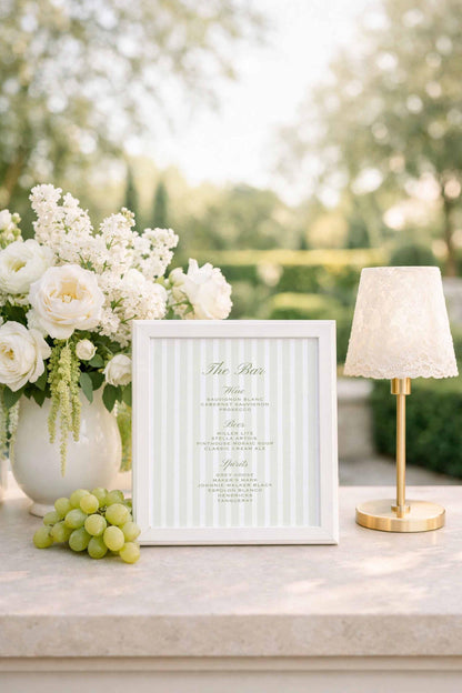 a white framed menu with a green stripe, placed on a table next to a vase of white flowers and a small lamp.
