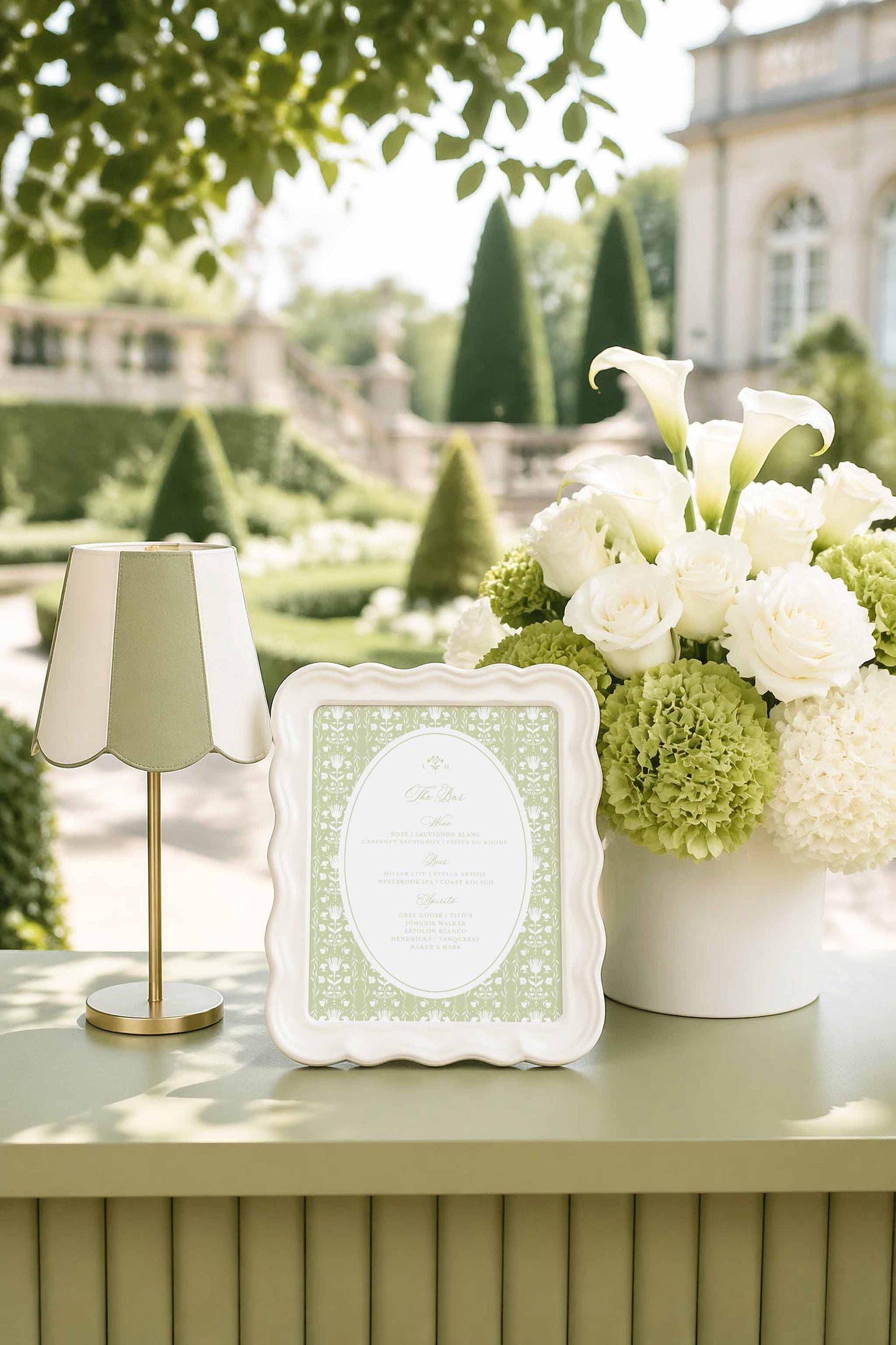 a table with a white vase filled with white flowers, a framed green and white menu, and a lamp. The table is set against a backdrop of a well-manicured garden with trees and a building.
