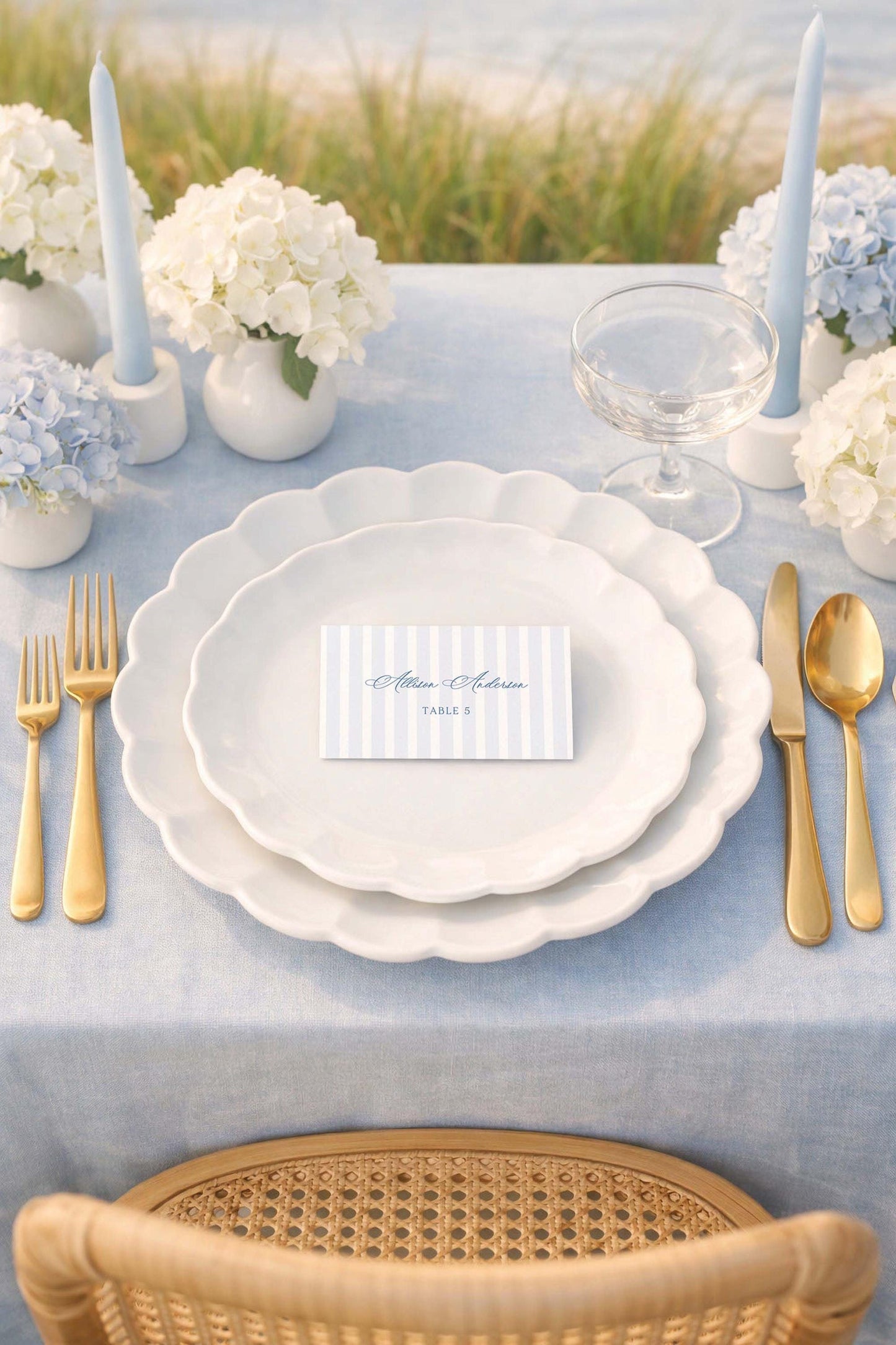 a beautifully set table with a blue tablecloth, white plates, and gold cutlery. There are two white vases with flowers, and a card with the name 'Amanda Anderson' on it. The table is set for a formal dining experience, with a wicker chair in the foreground and a serene outdoor view in the background.