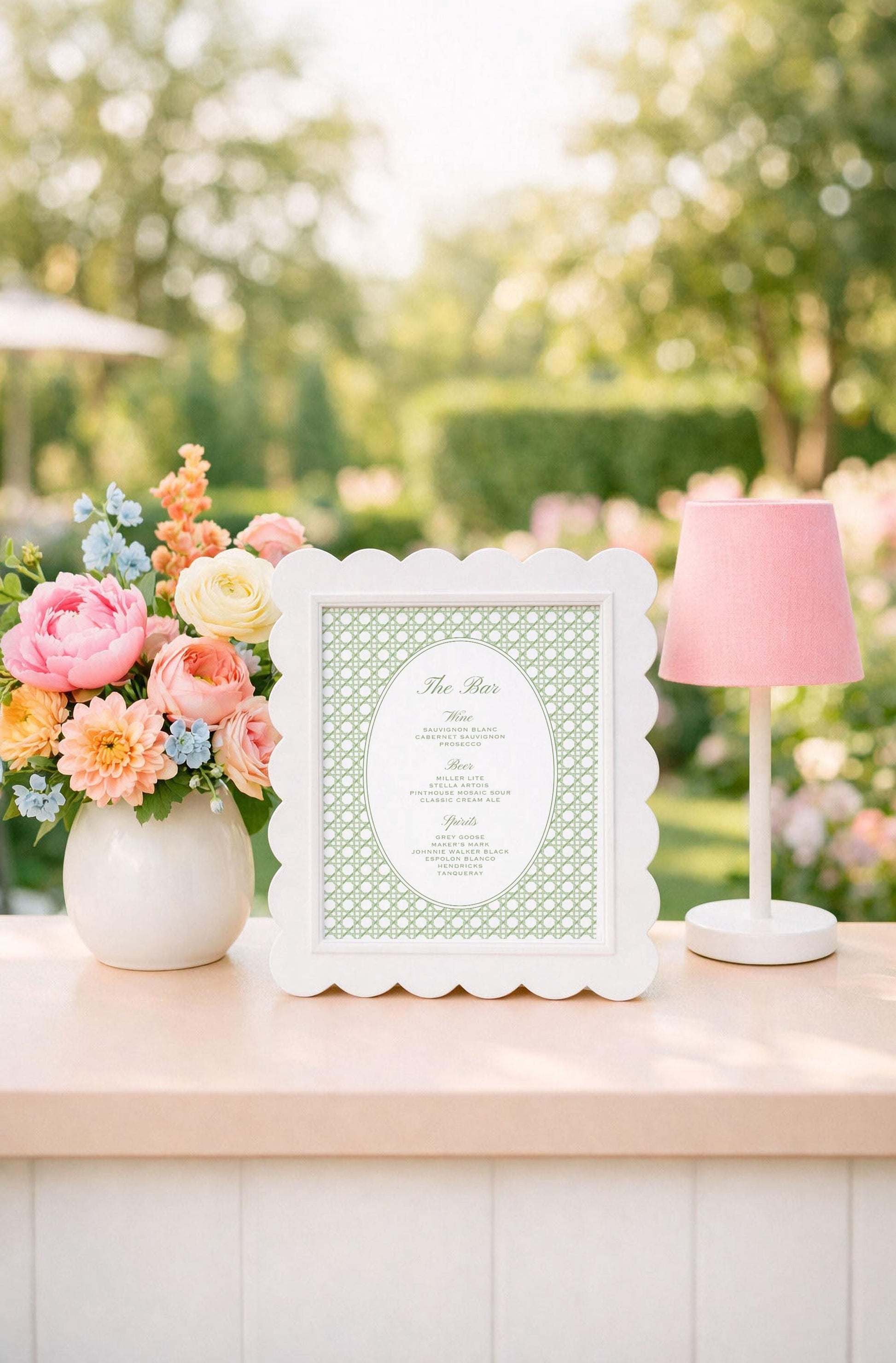 a white frame with a green pattern, a vase of colorful flowers, and a pink lamp on a table, all set against a backdrop of trees and greenery.
