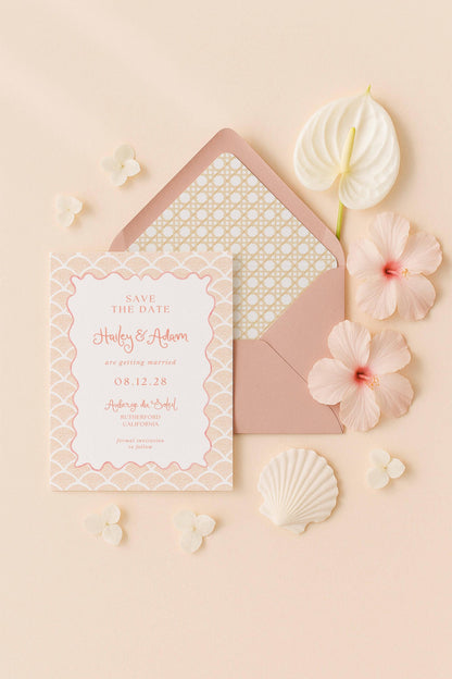 A pink wedding invitation card with a floral pattern, surrounded by pink flowers, shells, and a white hibiscus flower.